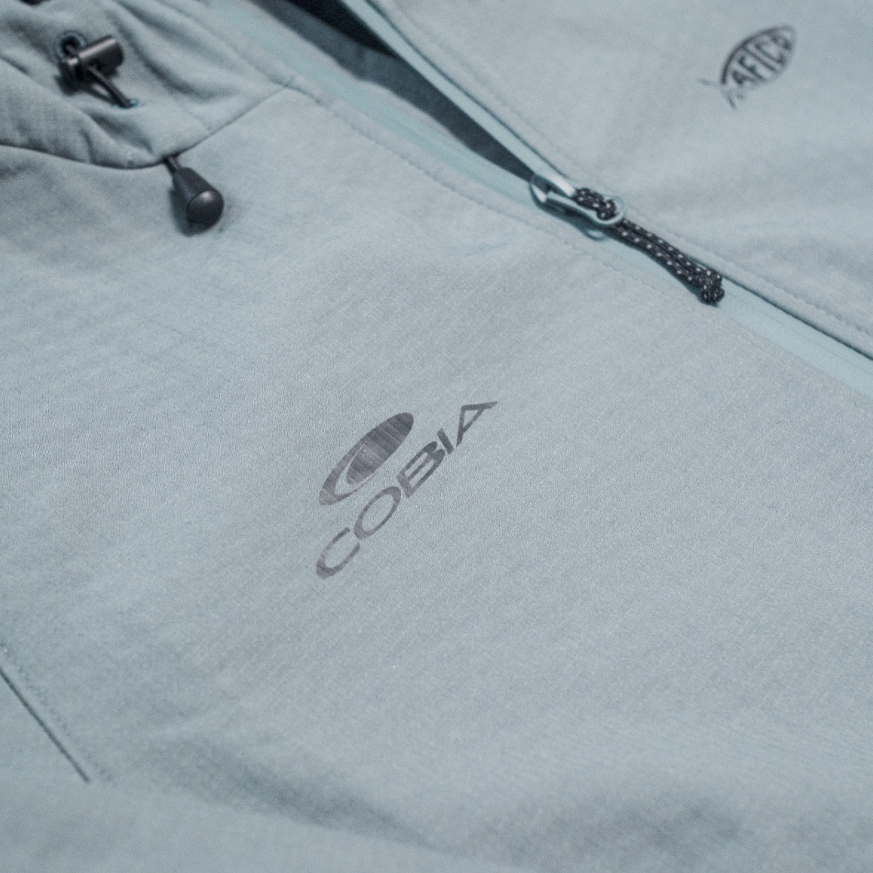 Close-up of a light gray jacket featuring the COBIA logo in grey on the chest.